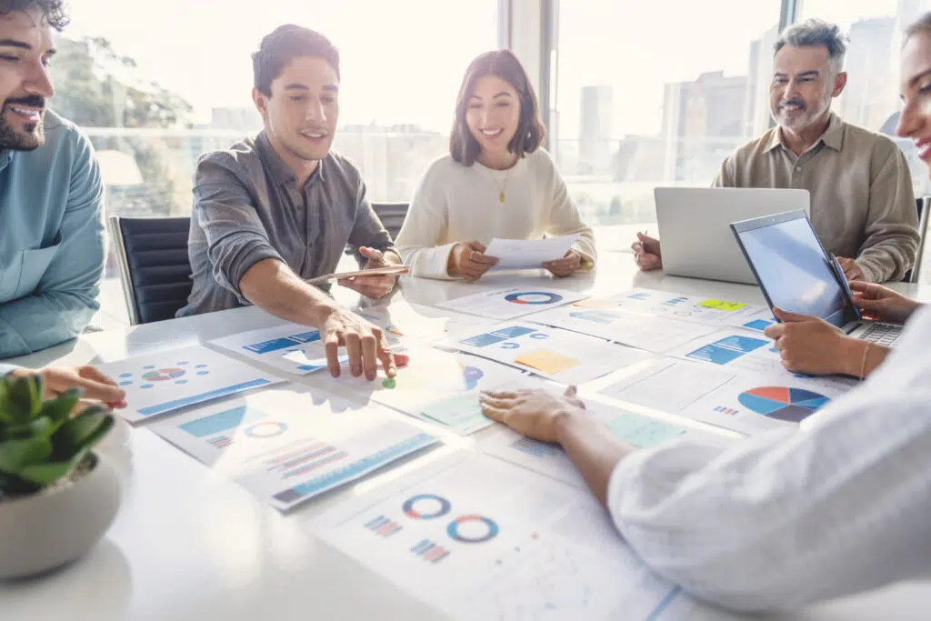 ulti racial diverse group of people working with Paperwork on a board room table at a business presentation or seminar. The documents have financial or marketing figures, graphs and charts on them. People are pointing to different documents. There are laptops and digital tablets on the table